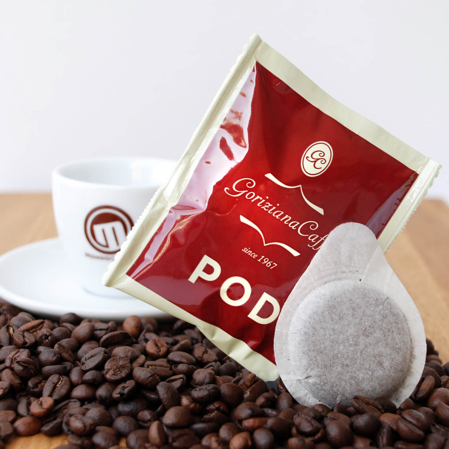 Die Abbildung zeigt eine Schachtel Torrefazione Goriziana Espresso ESE Pads (72 x 7 g) mit Kaffeepads, Verpackung, Bohnen und einer weißen Tasse mit Untertasse auf einer Holzfläche.